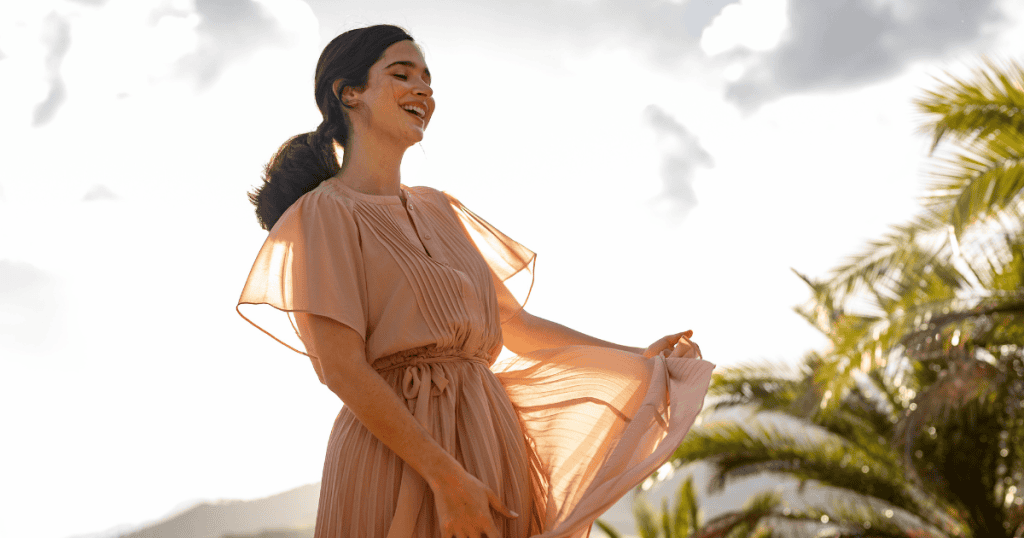 A smiling woman in a flowing peach dress looks up at the sunny sky, embracing the sense of relief found in prioritizing her own peace.