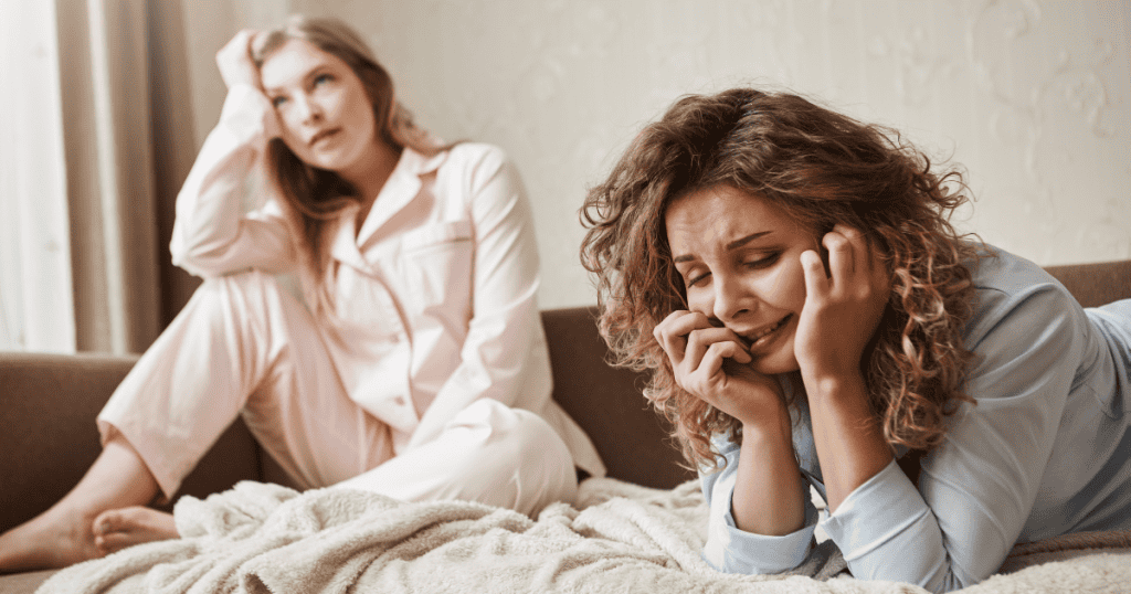 A woman lies weeping in the foreground while another sits indifferently behind her, visibly showing that the visible pain elicits zero emotional response.