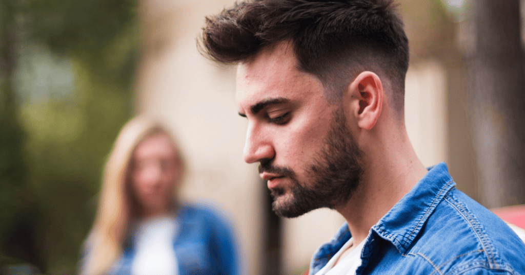 A man in a denim shirt looks down with a somber expression while a woman stands blurred in the background, capturing a moment of mental withdrawal from a shared space.