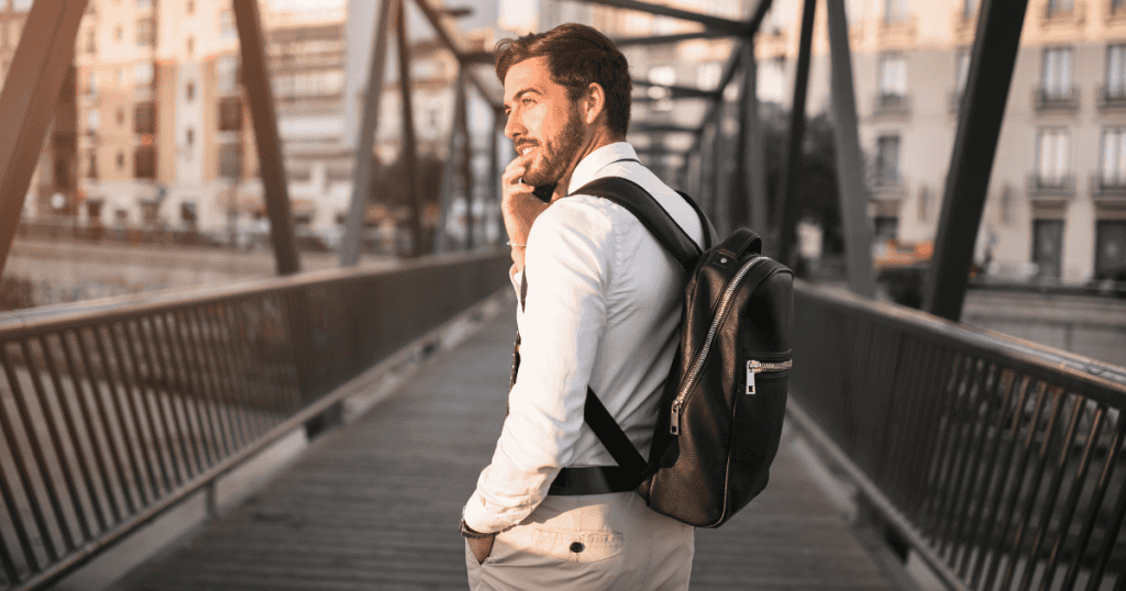 A bearded man with a backpack walks across a bridge while smiling into his phone, embodying the casual detachment of someone moving on to a new chapter without a backward glance.