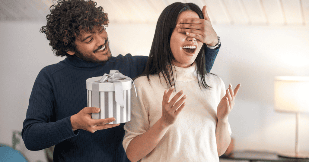 A smiling man covers a woman’s eyes while holding a gift behind her, illustrating how narcissists use gifts to distract, control, or buy silence instead of addressing real issues.