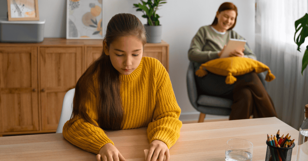 A girl focuses intently on her desk work while a woman watches from the background, reflecting a dynamic where a childโs efforts serve another's agenda.