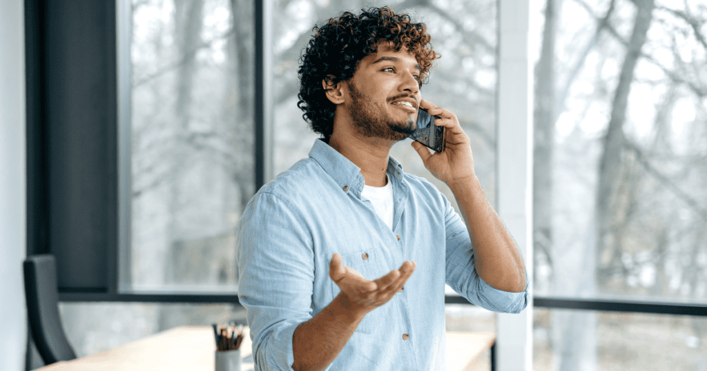 A man with curly hair smiles while talking on a smartphone and gesturing with an open palm, portraying a curated persona that deflects any sense of personal fault.