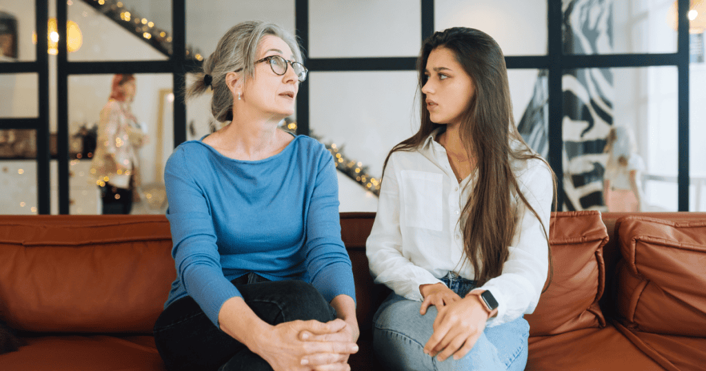 An older woman in a blue sweater speaks to a younger woman on a leather sofa, their forced proximity highlighting a disconnect in emotional sincerity.
