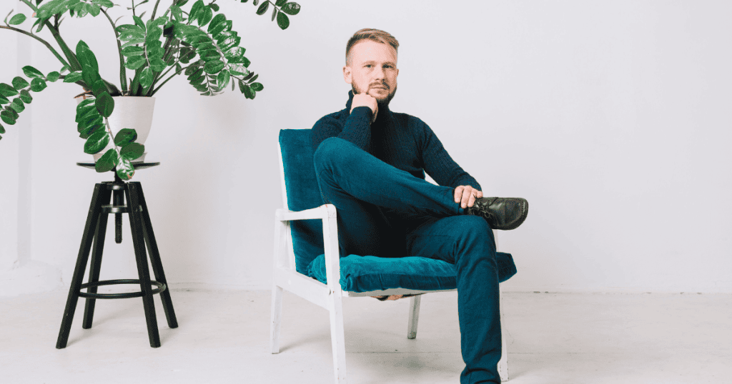 A man sits in a teal armchair with a neutral, expectant gaze; his rigid posture suggests a silent standard he expects others to instinctively meet.