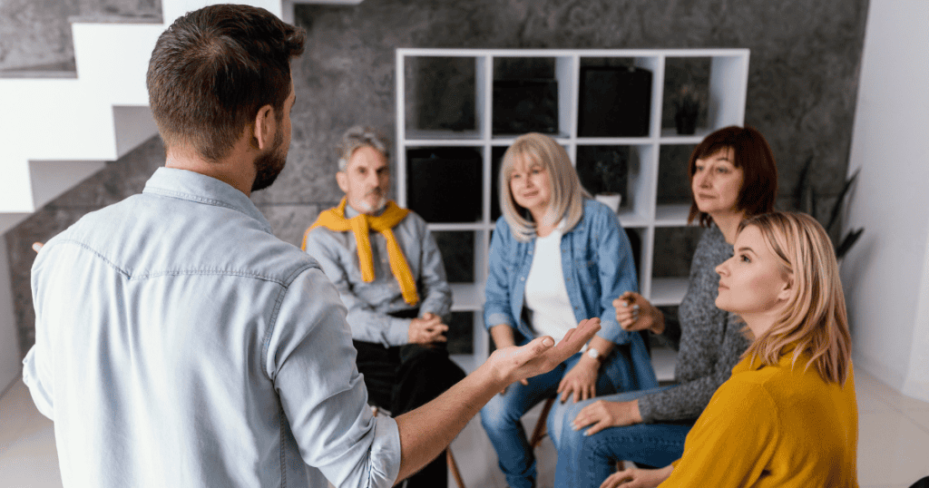 A man speaks to a seated group of people in a minimalist room, capturing a moment where difficult truths are finally being voiced to those who stayed.