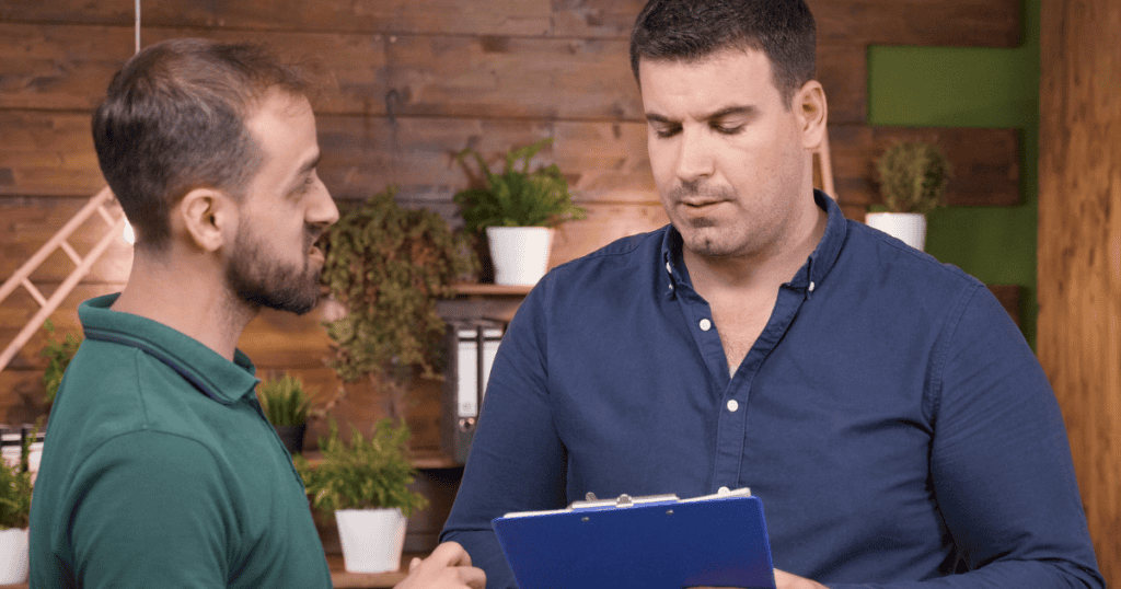 A man in a green shirt listens intently to a colleague holding a clipboard, a submissive stance that inadvertently strengthens the existing hierarchy between them.