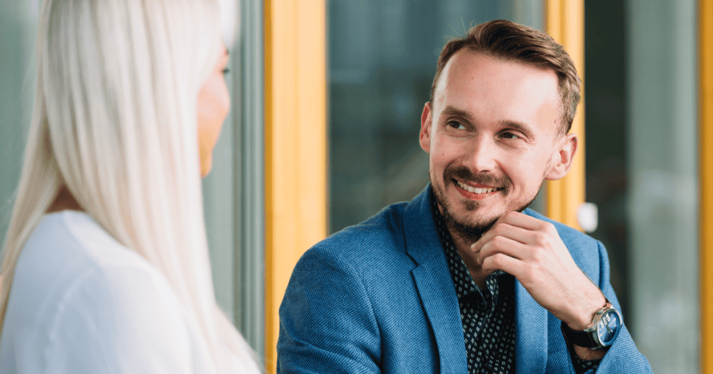 A man in a blue blazer leans forward with a charming, charismatic grin toward a woman, performing a level of engagement he selectively denies to those closest to him.