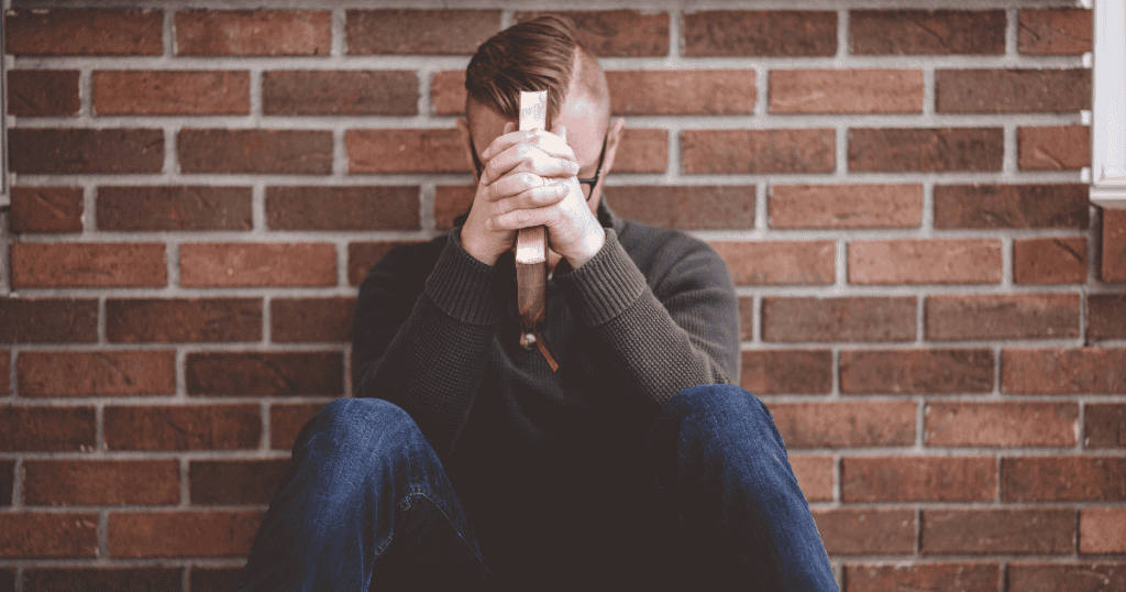 A man sits against a brick wall with his head bowed and hands tightly clasped over a book, reflecting the internal collapse that happens when a carefully constructed persona is finally breached.