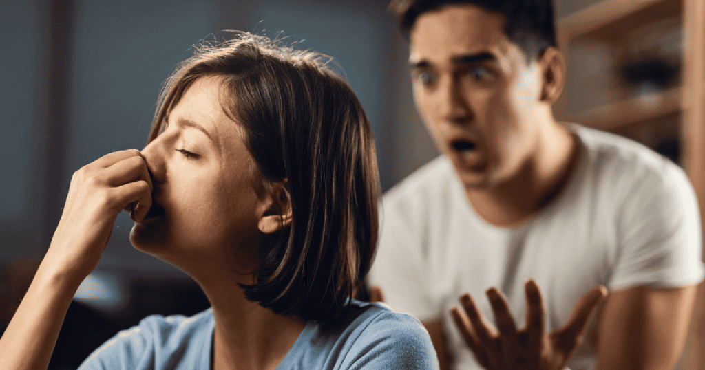 An agitated man gestures aggressively toward a woman rubbing her temples, his explosive reaction turning her moment of pain into a theatrical confrontation.