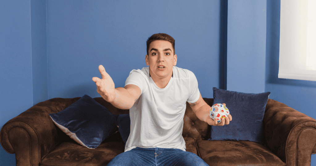 A man on a couch reaches out with an open palm while clutching a ball; his frustrated expression suggests a game where the goalposts are constantly moving.