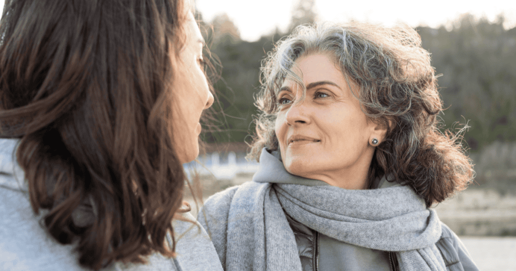 An older woman looks at another woman with a composed, almost gentle expression, illustrating the unsettling contrast of calm, controlled cruelty hidden beneath a steady gaze.