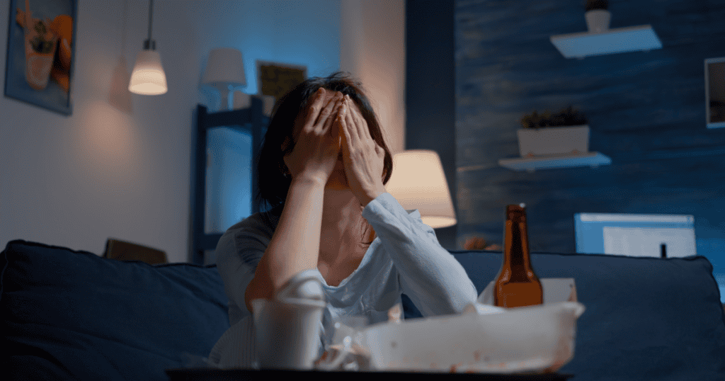 A woman sits on a couch at night covering her face in exhaustion, with takeout containers and a bottle nearby, illustrating how survival mode and emotional burnout become normal in a narcissistic relationship.