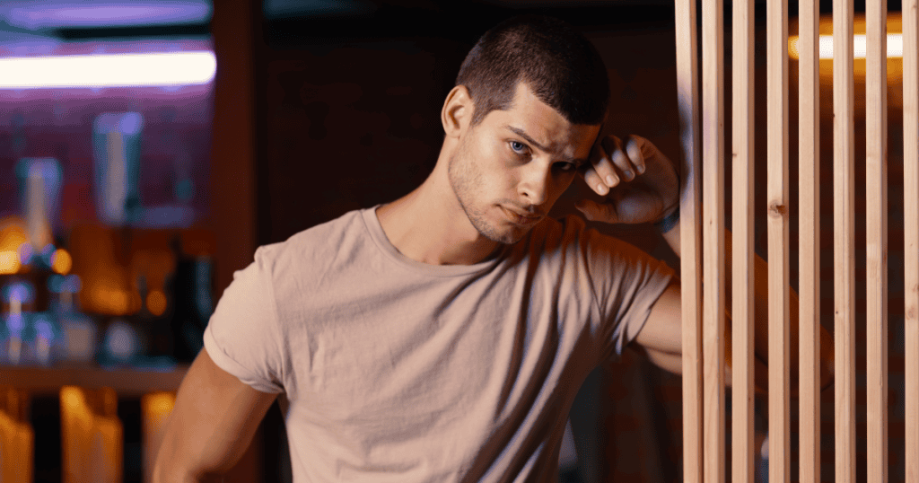 A man leans against a wooden divider with a brooding, intense expression, projecting confidence on the surface while subtly revealing the insecurity and fragility beneath the grandiose exterior.