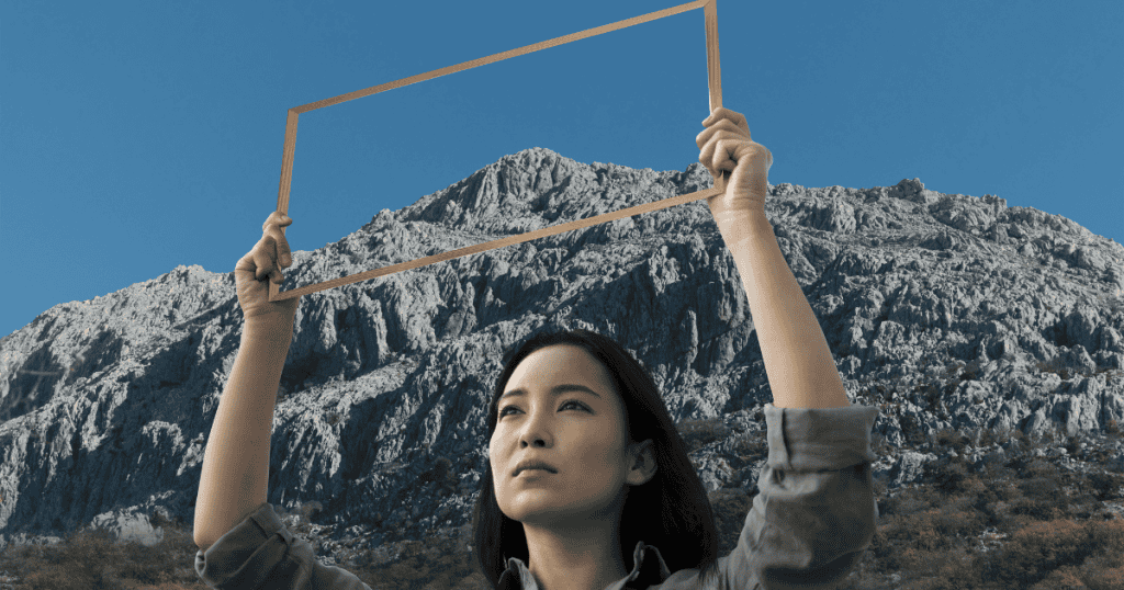 A woman holds an empty wooden frame up against a rugged mountain landscape, symbolizing the intentional act of choosing a new perspective to inhabit.