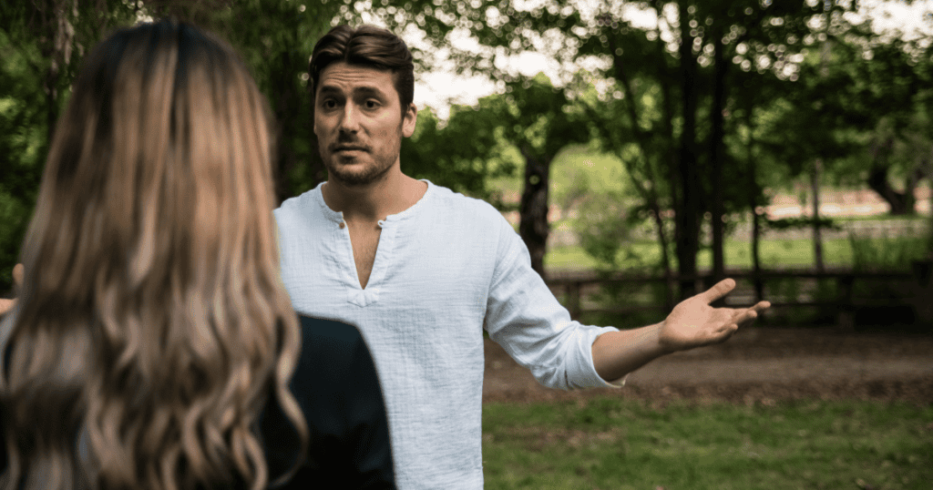 A man with open, defensive hands speaks to a woman outdoors, reflecting how a narcissist avoids admitting fault and deflects responsibility instead of taking accountability.