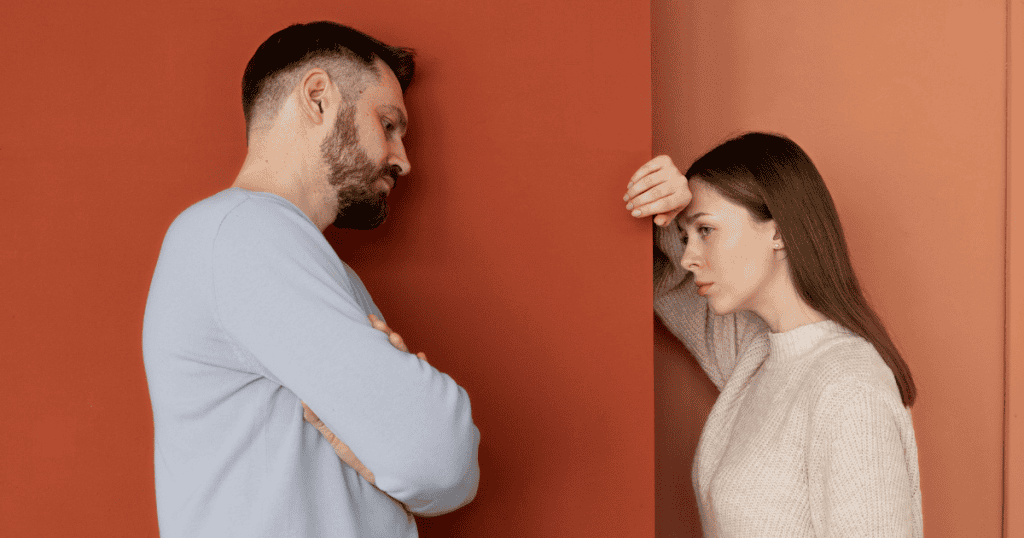 A man and woman stand on opposite sides of a thick red wall, capturing a moment where physical proximity cannot bridge a fundamental emotional divide.
