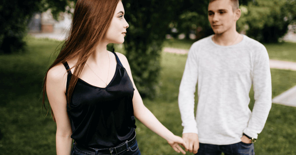 A woman walks away while still holding the hand of a man behind her, portraying a physical disconnect that hasnโt yet reached the fingertips.