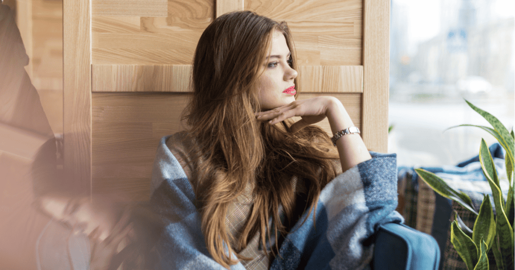 A pensive woman sits by a window with a ghostly reflection beside her, representing the lingering echoes of a cycle she is only beginning to name.