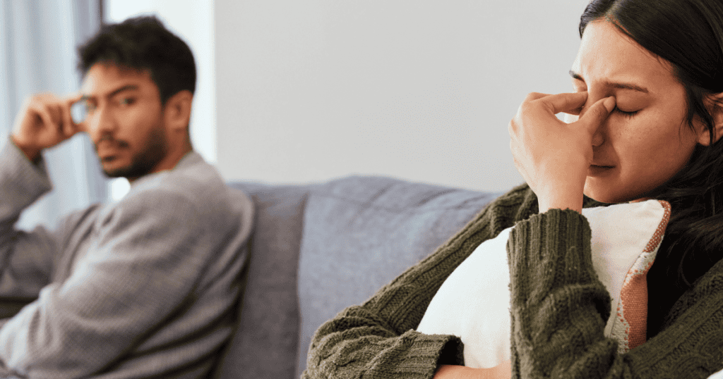 A woman sits curled up on a sofa in deep distress while a man watches coldly from the background, his stoic gaze framing her weakness as a calculated provocation.