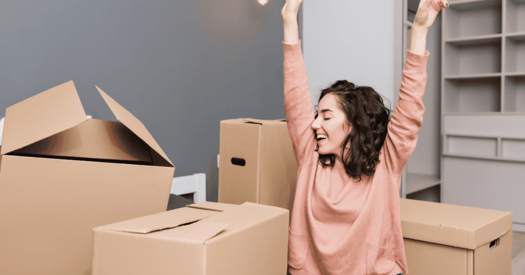 A woman cheers with her arms raised high among several packed cardboard boxes, celebrating the beginning of a story she is finally writing for herself.