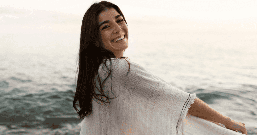 A woman looks back over her shoulder with a radiant, liberated smile at the beach, marking the moment she reclaimed her worth from someone else's influence.