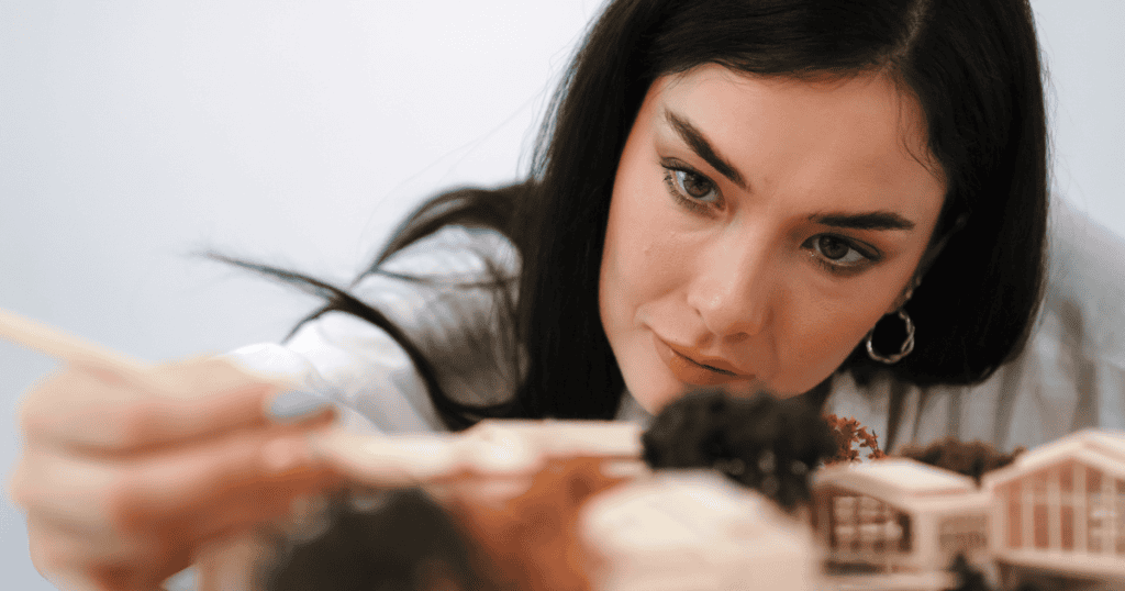 A woman meticulously adjusts a miniature architectural model with a small tool, focusing on the slow and precise labor of assembling a new foundation.
