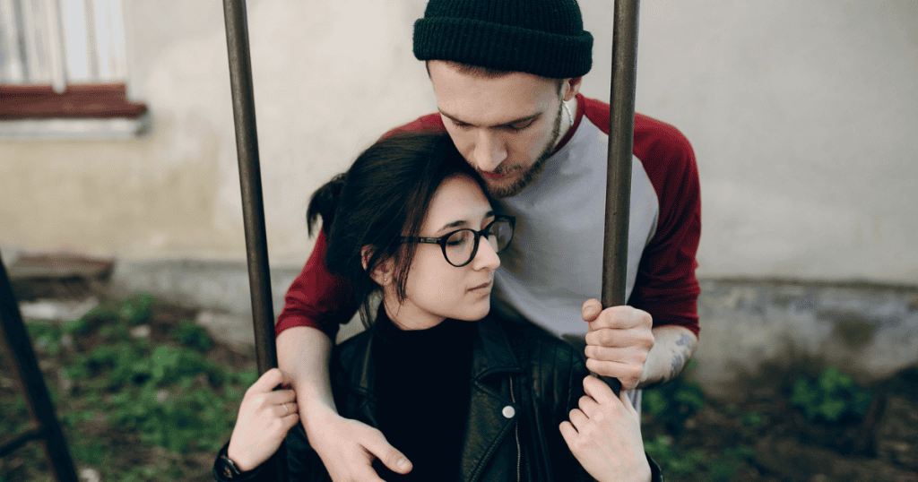 A man stands behind a seated woman and grips the metal poles of her swing, illustrating a protective gesture that feels more like an inescapable enclosure.
