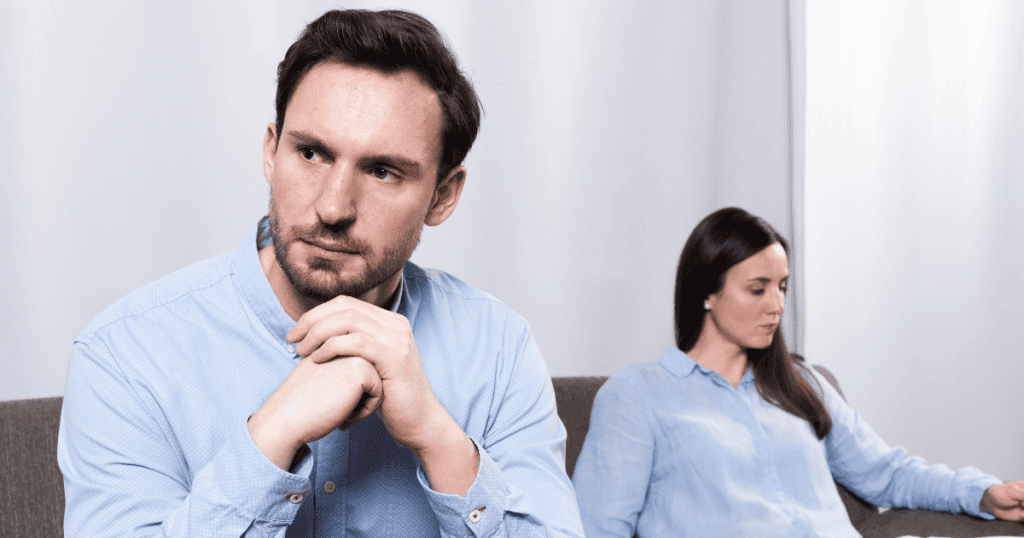 A man sits in the foreground with a distant, stony gaze while a woman remains blurred in the background, illustrating a deep emotional and physical disconnect.
