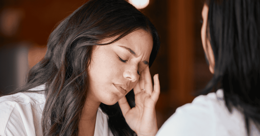 A woman sits with her eyes closed and a hand to her temple in a quiet room, showing the exhaustion of searching for an ending that cannot be granted by someone else.