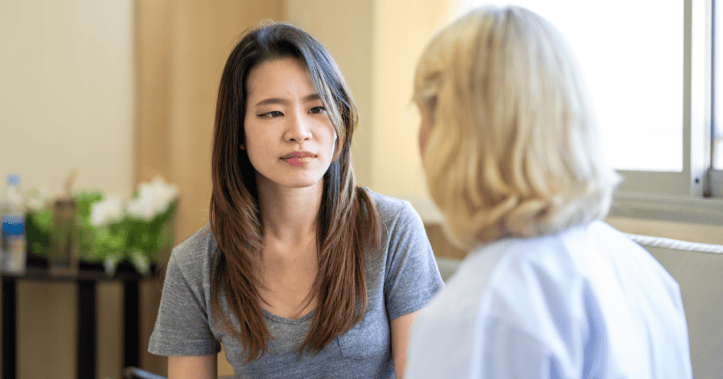 A woman sits quietly facing another person in conversation, her guarded expression reflecting how narcissistic dynamics silence you and make honest self-expression feel unsafe.