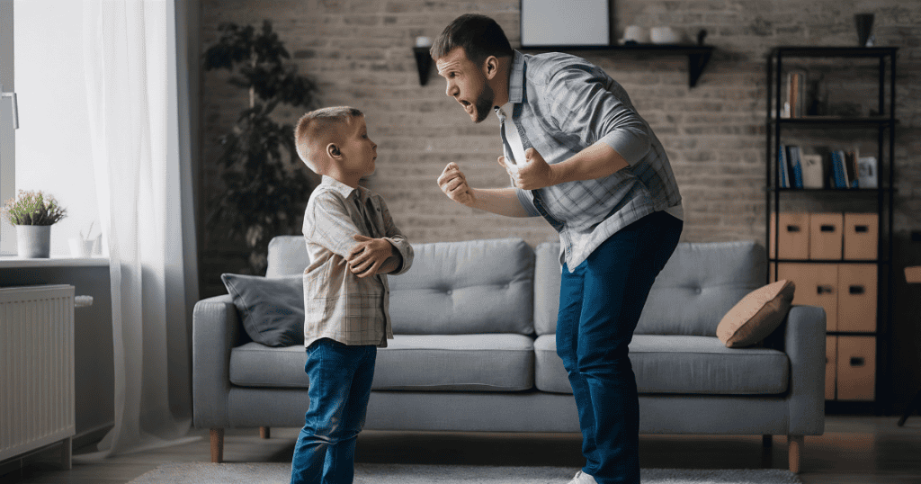 A young boy stands with his arms crossed and a stoic expression while a man gestures intensely nearby, showcasing the strength of a child who remains unmoved by external pressure.