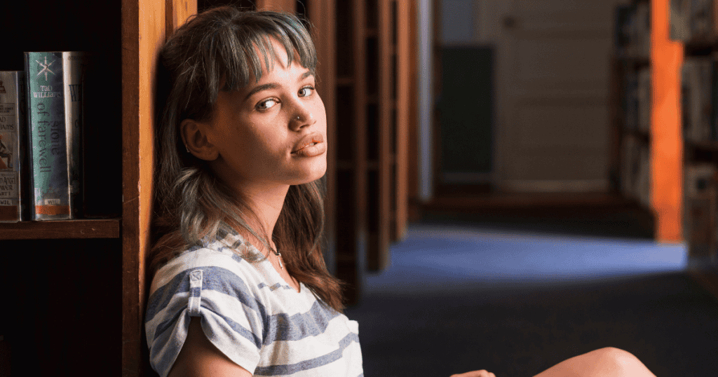 A young woman with green-tinted hair leans against a wooden bookshelf with a vacant, detached expression, capturing a moment of calculated indifference.