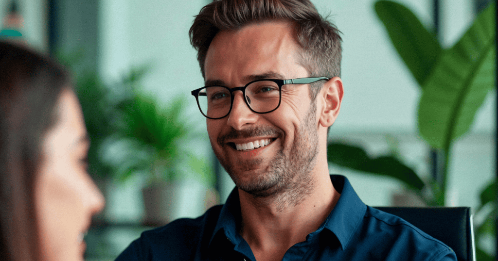 A man in glasses offers a sharp, toothy grin toward a colleague, his charming expression masking a pointed intent directed at his companion.