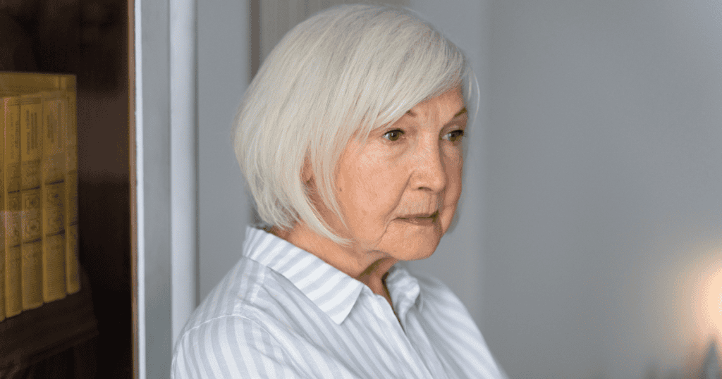 An older woman with white hair gazes pensively out of frame while standing near a bookshelf, capturing a somber moment of unavoidable internal reflection.