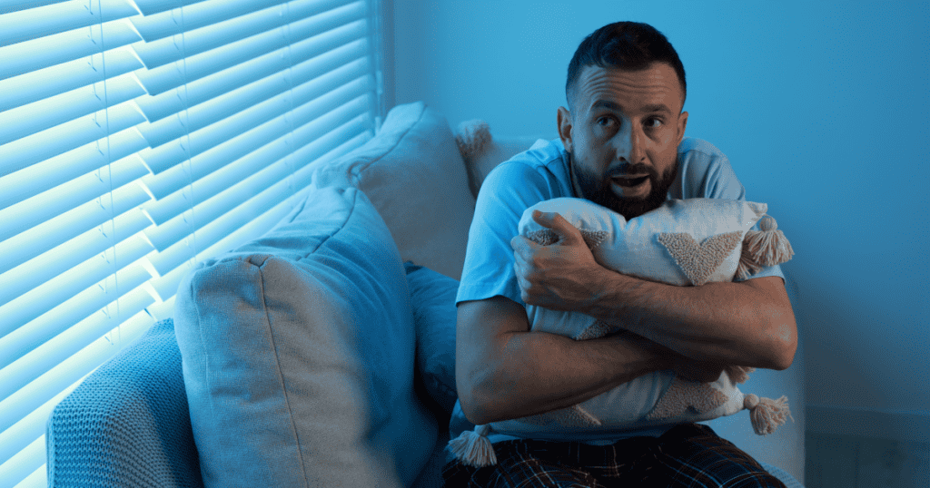 A tense man sits on a couch clutching a pillow defensively, his anxious expression reflecting the deep fear and insecurity narcissists hide and refuse to admit, even to themselves.
