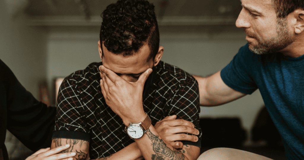 A distressed man covers his face while another person places a hand on his shoulder, illustrating how a narcissist adopts a victim role to gain sympathy and subtly reshape the narrative.