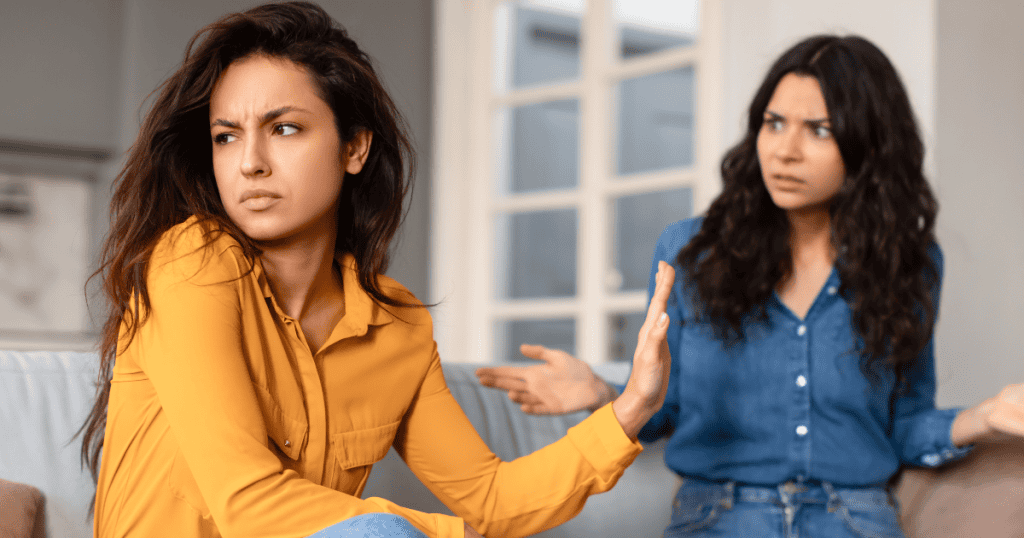 Two women sit on a sofa during a tense disagreement, one turning away with a dismissive hand gesture as if certain phrases are no longer up for discussion.