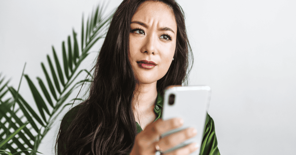 A woman in a green shirt looks away from her smartphone with a skeptical, furrowed brow, capturing the uneasy moment when external validation seems to slip away.