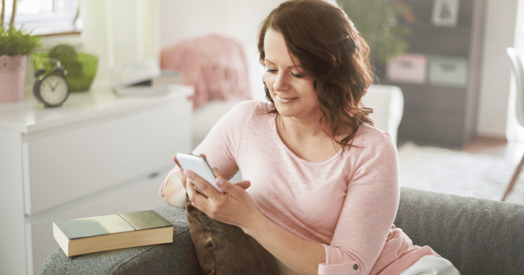 A woman sits comfortably on a sofa smiling at her phone screen, highlighting the quiet satisfaction found in constant digital engagement and attention.