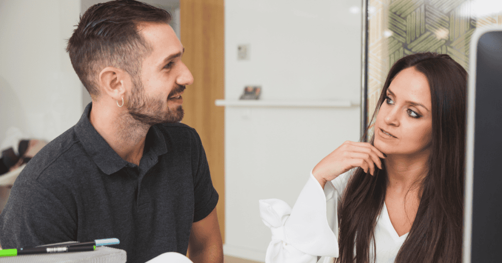 A woman listens intently to a man speaking at a desk, her analytical expression suggesting she is evaluating his words for something other than connection.