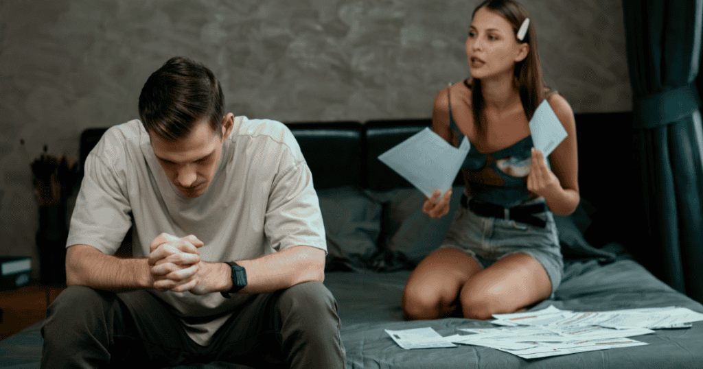 A man sits dejectedly on a bed while a woman gestures with papers, capturing a moment of heavy confrontation over broken trust.