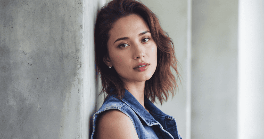 A woman with wavy brown hair leans calmly against a concrete wall while looking directly at the camera, her steady gaze and relaxed posture conveying a quiet sense of closure and emotional peace.