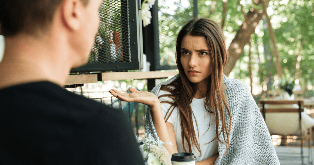 A woman looks at a man with a skeptical, questioning hand gesture during a conversation, marking the moment when a partner's behavior finally makes sense.