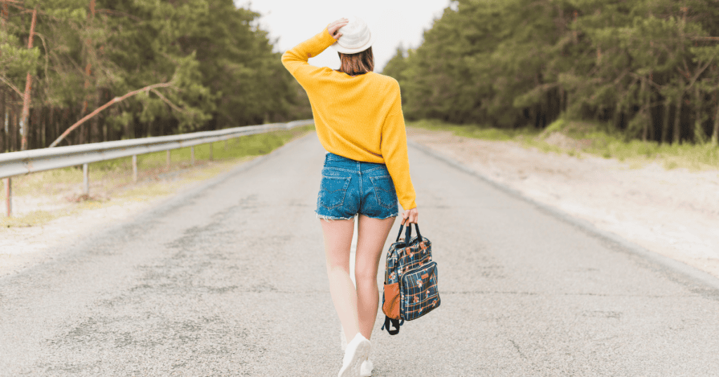 A woman in a yellow sweater and denim shorts walks down the center of an empty road into a forest, marking a solitary journey toward personal independence.