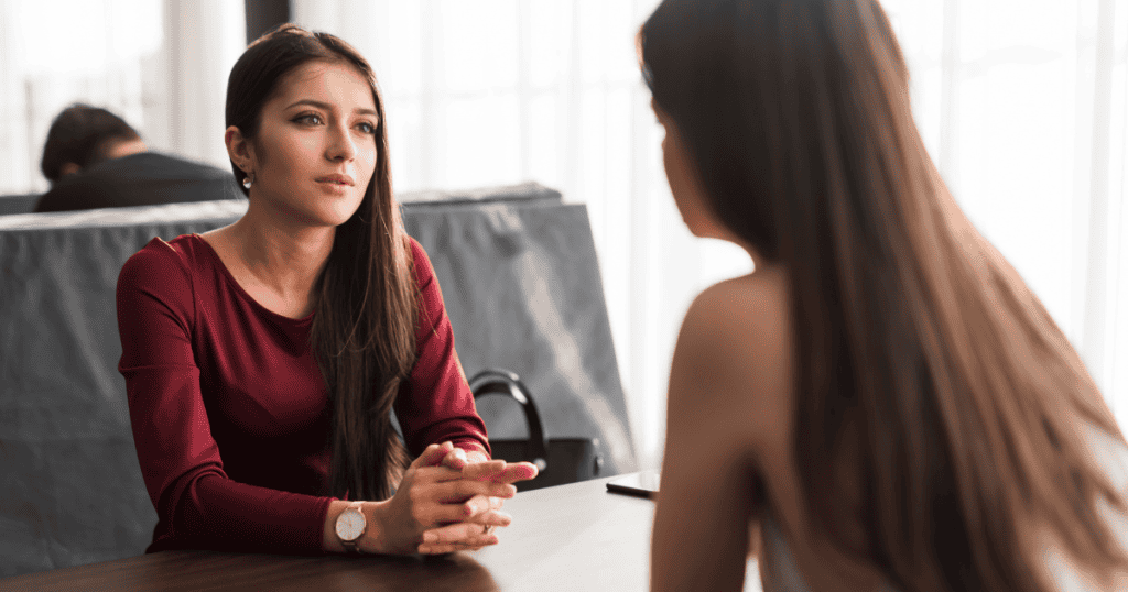 A young woman sits at a table with her hands calmly clasped while maintaining a steady gaze toward another person, capturing the grounded composure needed to maintain personal boundaries during a difficult exchange.
