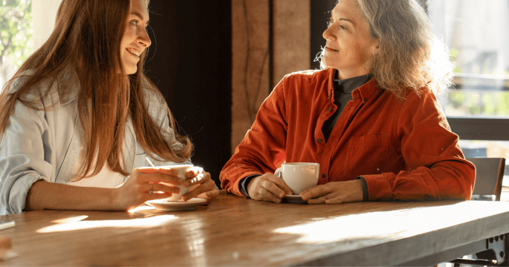 Two women sit across from each other at a sunlit cafe table with coffee, their polite smiles masking the underlying social evaluation taking place between them.