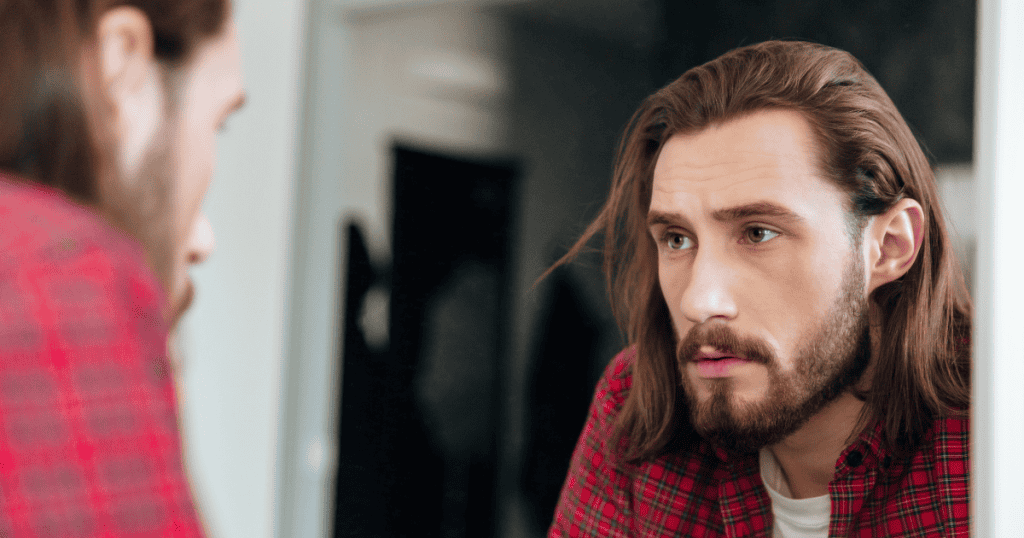 A man with long hair stares intensely at his own reflection in a bathroom mirror, capturing a moment of profound self-scrutiny and the complex layers revealed by psychological observation.