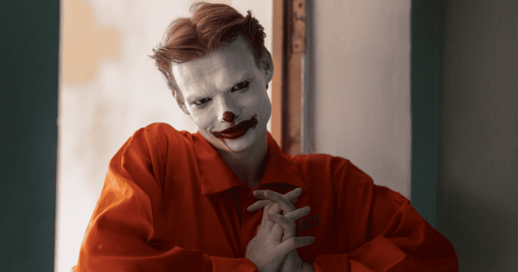 A man with a calculated, unsettling smile and exaggerated appearance stands with hands clasped, symbolizing the manipulative charm narcissists use, and the importance of recognizing it so you’re never fooled again.