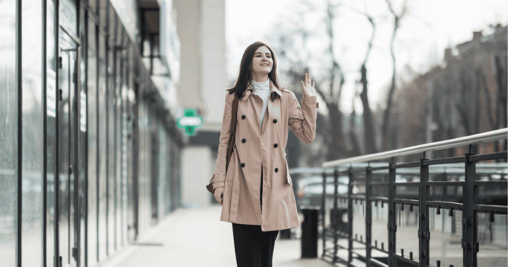 A woman in a tan trench coat walks down a city sidewalk while waving goodbye with a smile, representing the liberating act of physically and emotionally exiting a draining dynamic.